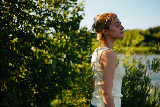 A woman stands still near a body of water, lost in thought while enjoying the warmth of the sun. The lush greenery surrounds her, creating a serene backdrop for this moment