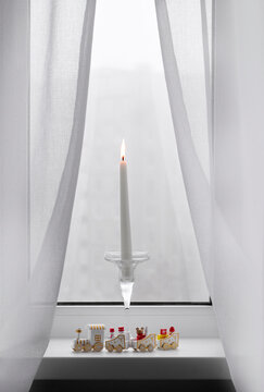 A lit white candle in a glass holder and a Christmas toy train decoration on a window sill covered by light curtains