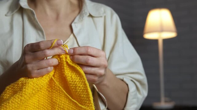 Woman's hands knitting yellow yarn indoors. Young woman's hands knitting with needles using bright yellow wool yarn. Cozy and calming home hobby creating handmade warm clothes