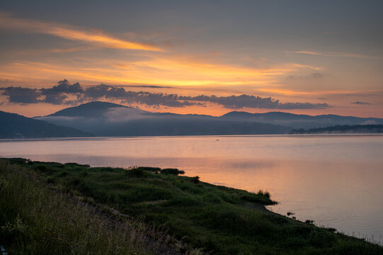 A glowing orange and golden sunrise sky reflects on a calm mountain lake with grassy shore and misty hills in the distance. Taken by Żywiec Lake.