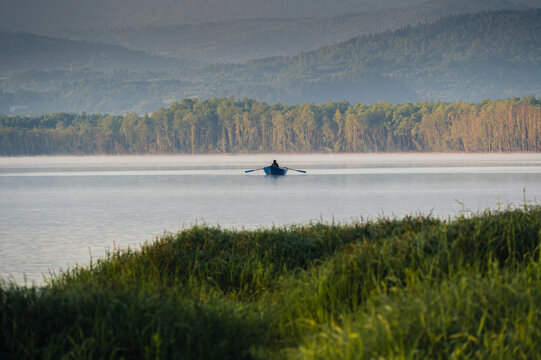 Small rowing boat on a calm misty lake with green reeds, forested hills and mountains in soft morning light.
