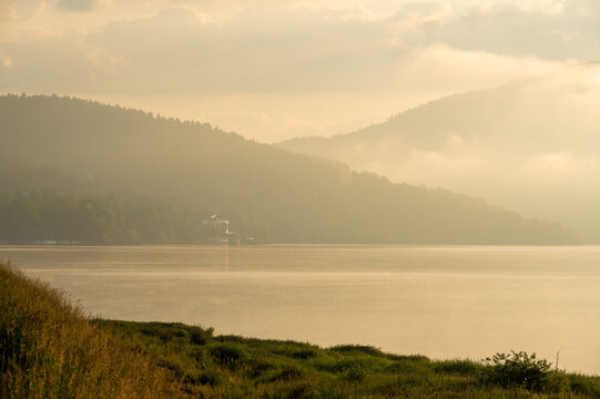 Atmospheric golden morning light over a misty lake with layered hills, calm water and soft summer haze.