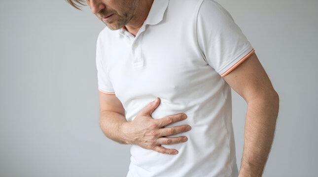 A middle aged man wearing a crisp white polo shirt experiences sudden sharp abdominal pain while holding his stomach with his right hand due to severe indigestion or medical issues.