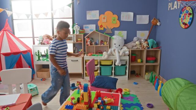 African american boy running and reaching with bare hand for a toy block amid scattered bricks in bright playroom building; playful curiosity.