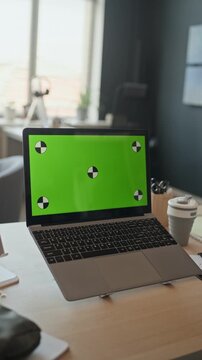 Vertical shot of laptop with green screen and tracking markers standing on wooden desk in contemporary office. Mockup display allowing easy integration of custom software or website designs
