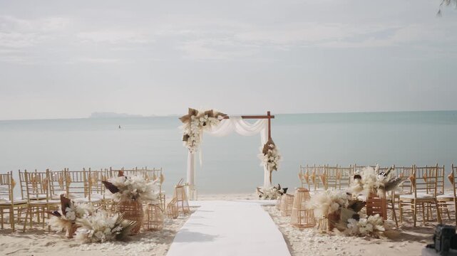 A stunning bohemian beach wedding ceremony setup on white sand, featuring a wooden arch adorned with pampas grass, white flowers, gold chiavari chairs, and a white aisle facing the ocean.