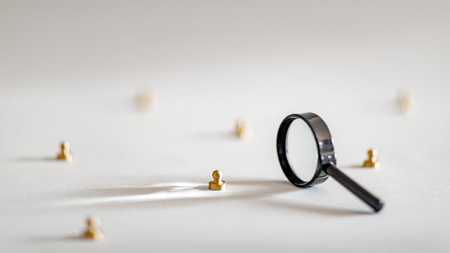 A black magnifying glass near small golden figures. Conceptual metaphor for human resources recruitment, headhunting, market audit, or finding a unique leader.