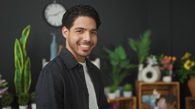Man smiling with potted plants and flowers in a studio, standing near wooden shelves, vases and a wall clock; warmth hospitality.