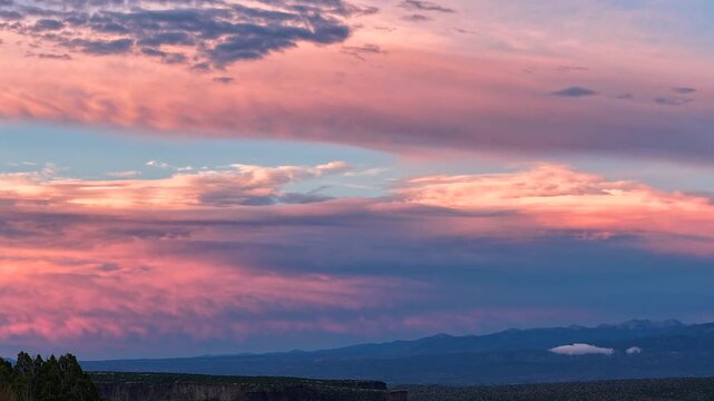 4K UHD drone timelapse of a surreal, thick curtain of clouds drifting over a dark mountain horizon. Vibrant sunset colors transition through the sky, illuminating the textured cloud layers.