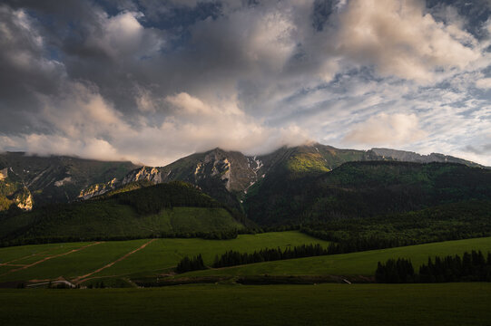 Wide dramatic view of the Belianske Tatras in Slovakia with Placliva Skala and Havran peaks in the center, low clouds covering the rocky summits, green forested slopes and moody light.