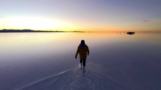 Back view of traveler walking through water reflection in Uyuni salt desert during golden hour