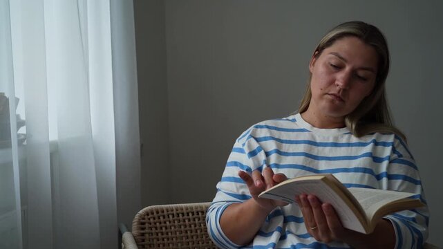 Casual reader in a striped top turns pages while sitting in a wicker chair near a bright window.