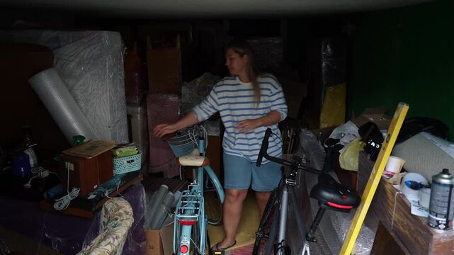 Adult woman squeezes between stacked furniture and bicycles in an overfilled garage, attempting to organize the chaotic storage space.