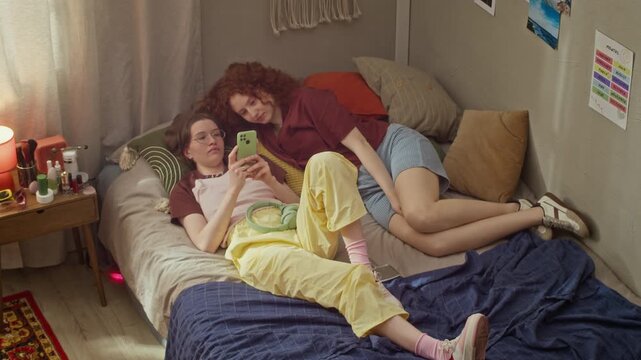 Caucasian girls lying on bed, scrolling on smartphone and laughing together. Footage is suitable for mobile application marketing and youth technology campaigns, high angle wide stab