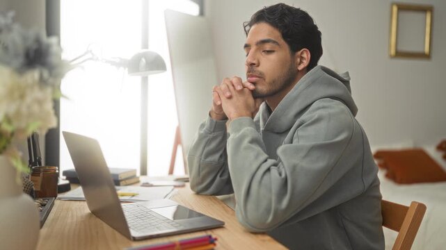 Man with laptop and hands clasped at desk in bedroom, leaning forward with eyes closed and focused expression while seated at a home workspace; pensive contemplation.