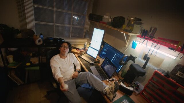 Portrait of young adult Asian male programmer sitting at desk with feet up, tossing ball while taking break from coding. Clip is ideal for tech recruitment and remote work lifestyle videos