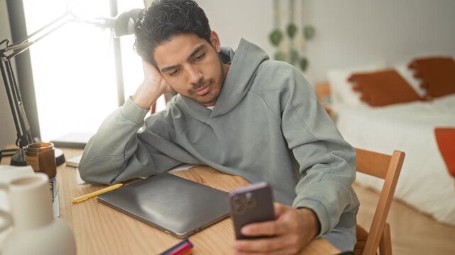 Man holds smartphone and leans head on hand over laptop at wooden desk with lamp and bed visible in studio; quiet contemplation.