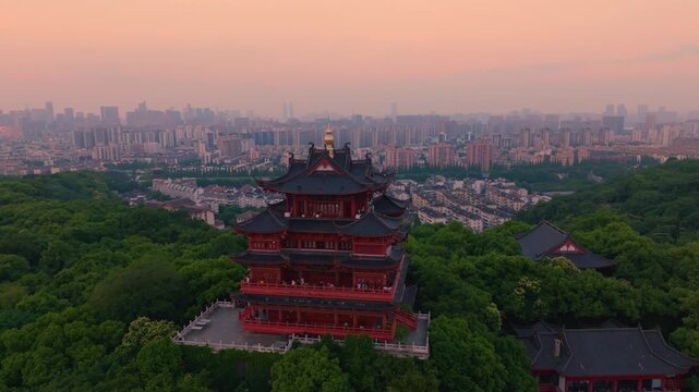 Aerial shot of Chenghuang Pavilion landmark of West Lake Hangzhou, traditional Chinese multi-story pavilion surrounded by green trees, distant city skyline under dusk pink sky, World Cultural and Natu