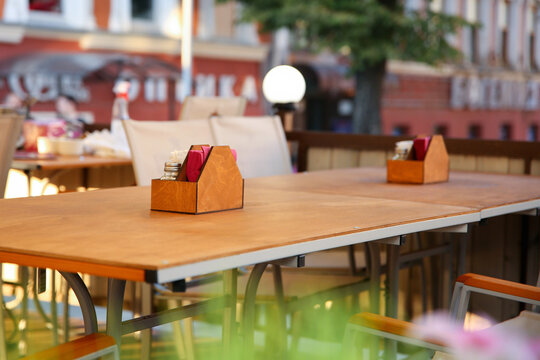 Empty wooden tables at a cozy outdoor cafe terrace. In focus is a wooden napkin holder. Soft blurred city background. Calm, inviting atmosphere of a warm summer day