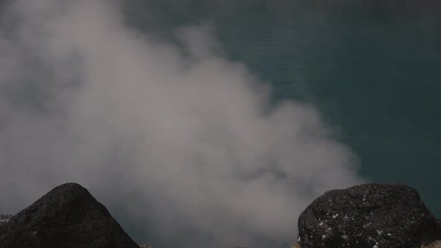 Thick white smoke and volcanic steam erupting from a crater, blowing rapidly in the strong wind. Dark rocky volcanic peaks stand in the foreground under a dramatic atmosphere. Beppu jigoku
