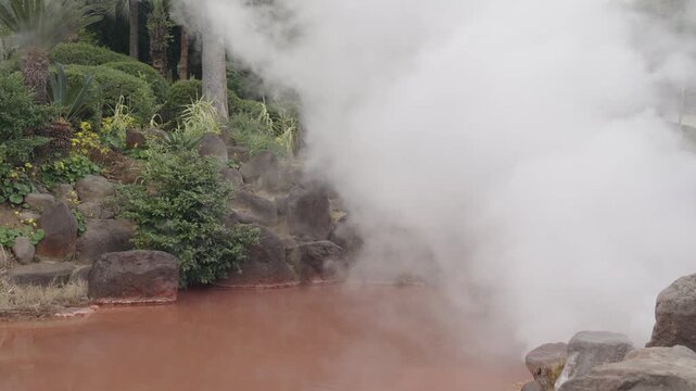 Thick hot steam and white smoke blowing across a reddish-brown geothermal hot spring pond by the wind. Scenic view of Chinoike Jigoku, the blood pond hell onsen landscape in Beppu, Japan.