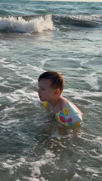 Happy boy in colorful arm floaties jumps and plays in sea waves on sunny beach with big smile. Carefree childhood summer sea play and safe swimming concept. Young child enjoys playing in ocean waves. 
