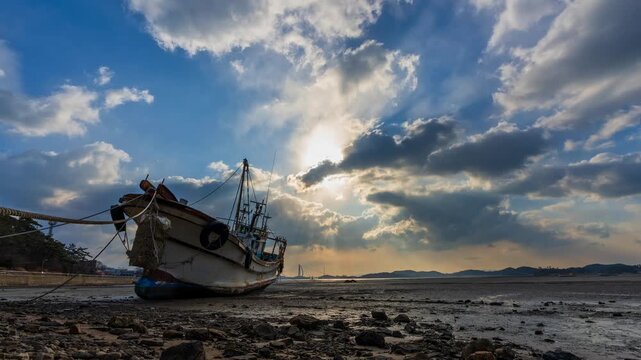 Timelapse of a fishing boat stranded on a muddy beach at low tide with dramatic sun rays piercing through moving clouds, coastal landscape view.