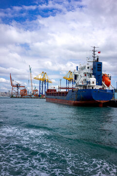 A blue cargo ship sits docked at Istanbul's industrial port alongside towering yellow and orange loading cranes under a dramatic cloudy sky over the Bosphorus.