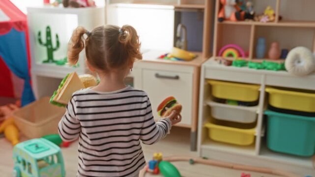 Girl holds toy sandwich and reaches for a toy microwave while playing at a pretend kitchen in a building playroom; playful bonding.