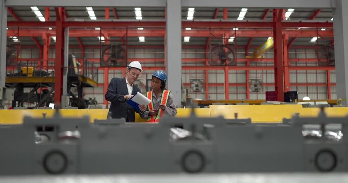 A senior manager and an African female worker in a safety vest use a digital tablet to inspect steel coils stored in a warehouse under an overhead crane hook discussing stock quality