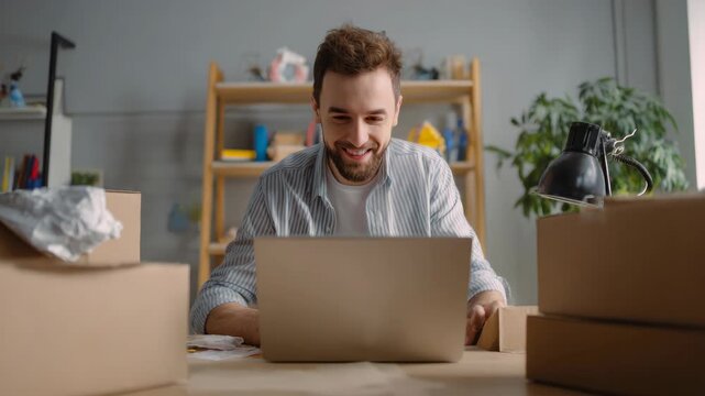 A smiling man with beard and brown hair using a laptop computer at work with cardboard boxes piled up behind him. He is wearing a striped shirt.