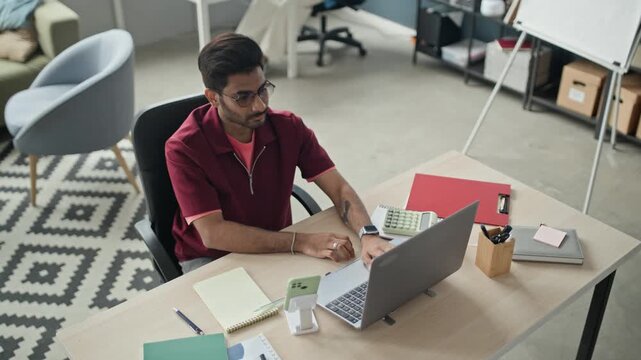 Overhead shot of Indian young adult man with glasses typing on laptop at office desk, pausing, coping with stress. Ideal for campaigns on employee burnout and mental health support