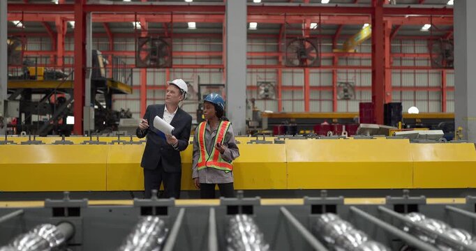 A senior manager and an African female worker in a safety vest use a digital tablet to inspect steel coils stored in a warehouse under an overhead crane hook discussing stock quality