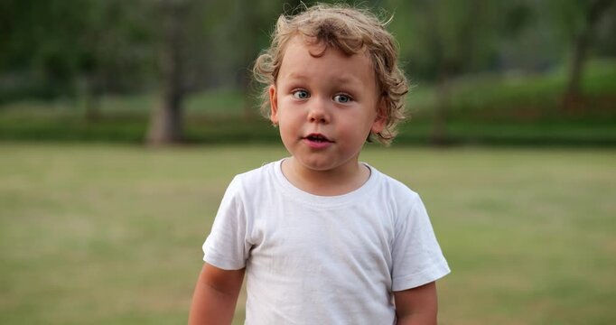 Wide-eyed, thoughtful baby boy stands in park at evening. He briefly responds, then glances away, lost in curiosity. Slow-motion portrait captures his innocent expression and gentle emotion
