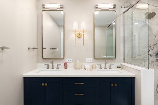 A bathroom with tan walls, gold and bronze fixtures, marble tiled shower, and a white marble countertop on a blue cabinet with gold hardware.