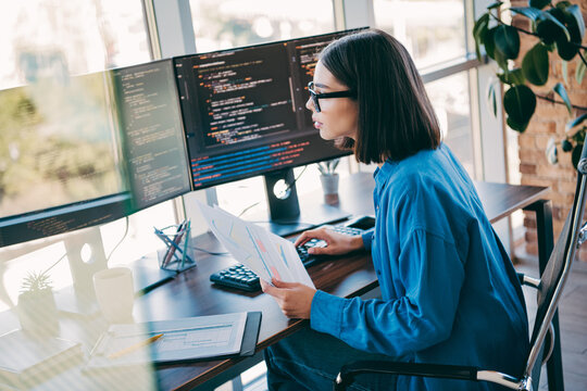 Female programmer at a sunny loft office coding on multiple monitors while reviewing documents