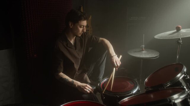 Portrait of Caucasian young male drummer sitting at drum kit in dark music studio, holding drumsticks and looking at camera. Footage for music education or band promotions