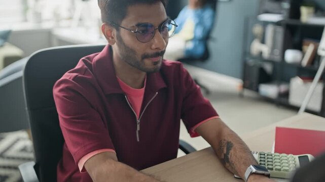 Tilt up of Indian young man with glasses typing on laptop, writing notes, and nodding his head and moving fists to music, sitting at desk. Suitable for corporate training, business strategy video