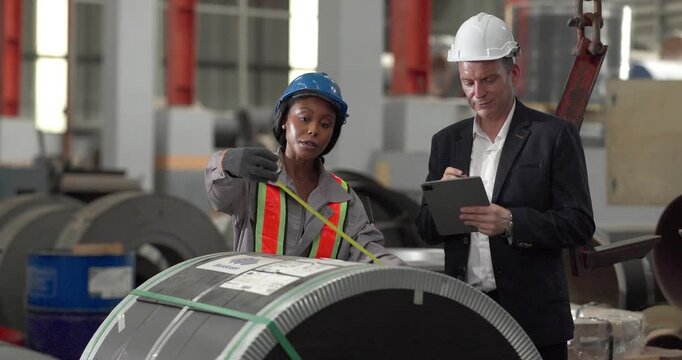 A senior manager and an African female worker in a safety vest use a digital tablet to inspect steel coils stored in a warehouse under an overhead crane hook discussing stock quality
