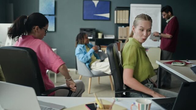 Waist up of two young adult Caucasian women spinning in office chairs, tossing apple and laughing with diverse male colleagues in background. Perfect for corporate culture and modern workplace videos