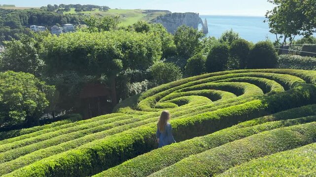 Cinematic horizontal shot of young woman in blue dress walking away through green spiral labyrinth towards Etretat white cliffs and ocean, France.