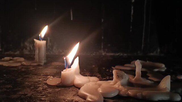 Close-up of burning white candles with melting wax and flickering flames against a dark background. Soft glowing candlelight with dramatic contrast. Spirituality, remembrance, relaxation, and peaceful