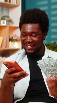 African American young man on home sofa counts cash and checks smartphone after online game win in living room. Black boy on home sofa compares amount and enjoys sports bet or casino success indoors