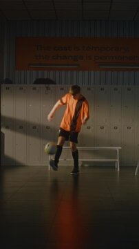 Talented Teenage Soccer Player in Orange and Black Kit Skillfully Juggling Football in Middle Empty Sports Dressing Room. Boy Maintains Focus, Control, Athletic Ability Between Lockers. Vertical Shot.