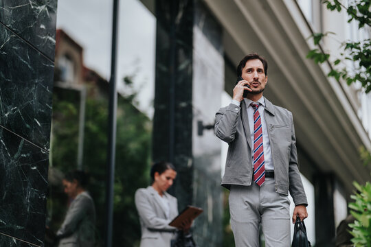 A businessman in a gray suit talks on his phone while walking outside an office building. A colleague stands in the background using a tablet, creating a professional urban work scene.