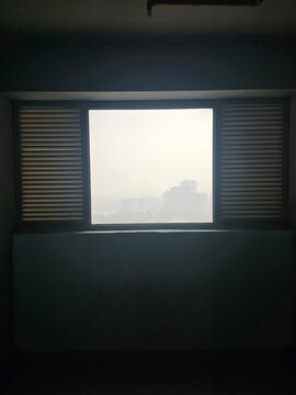 A view from a window framed by closed shutters, revealing a hazy urban landscape, creating a contemplative mood of isolation and urban life.
