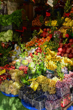 A vibrant, densely packed outdoor market stall showcases a large variety of seasonal fruits and greens. Clear plastic containers filled with strawberries, grapes, and plums