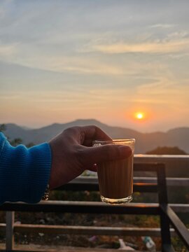 A hand holding a glass of tea against a backdrop of a serene sunset, with mountains in the distance, evoking a tranquil and peaceful evening atmosphere.