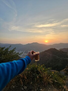 A hand holding a glass of tea against a stunning sunset backdrop, showcasing the beauty of nature and tranquility in the hills.