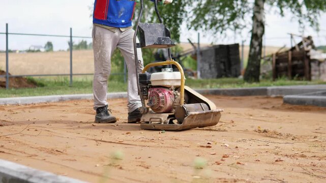 Worker guides plate compactor across sandy construction ground beside curb bordered worksite, showing surface leveling, earth compaction, and outdoor site preparation under soft